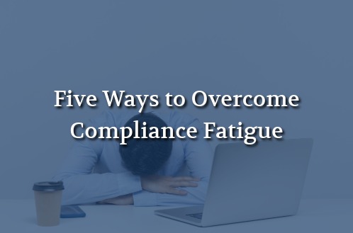 Fatigued professional resting their head on folded arms beside a laptop, illustrating workplace compliance fatigue.