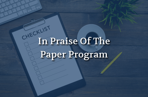 “In Praise Of The Paper Program” displayed over a checklist on a clipboard with a pencil and office workspace.