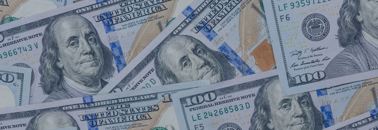 A scattered pile of U.S. one hundred dollar bills featuring Benjamin Franklin's portrait. The blue security ribbon and seal add vibrant color accents.