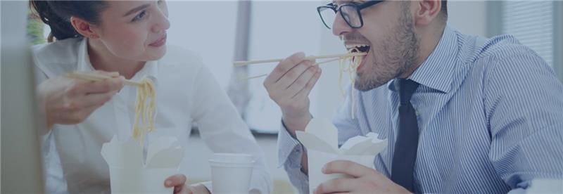 A woman and man in business attire enjoy noodles with chopsticks from takeout boxes in an office, sharing a light-hearted, casual lunch moment.