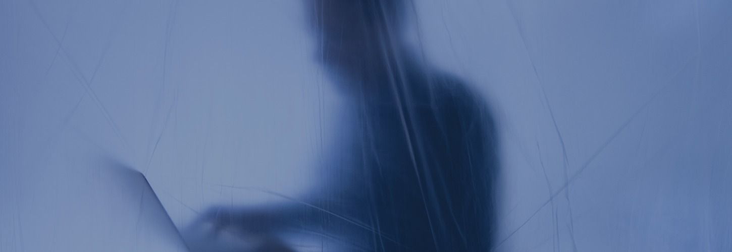 A blurred silhouette of a person typing on a laptop, seen through a blue, textured surface. The image conveys a mysterious, secretive mood.