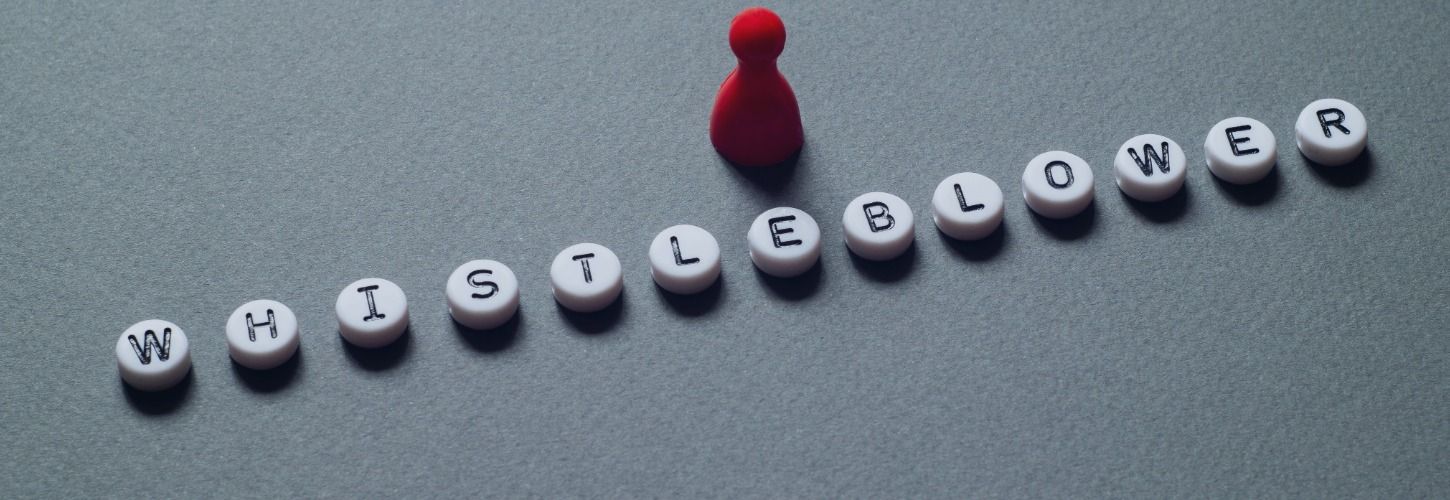 Alt text: "A red pawn figure stands above white lettered beads spelling 'WHISTLEBLOWER' on a gray background, conveying a theme of courage and vigilance."