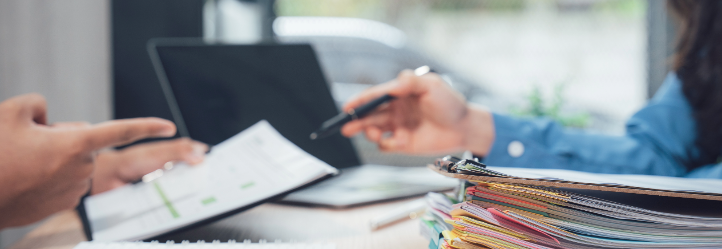 Photo of two people reviewing paperwork at a desk, with one person pointing to a document and another holding a pen near stacked folders and files, representing an investigation process or compliance review meeting.