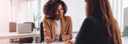Business consultant in a tan blazer speaking with a client across a desk in a bright office, holding a pen during a professional discussion.