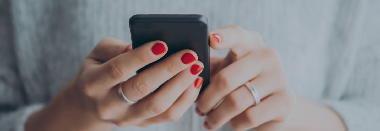 Hands with red nail polish holding a smartphone, typing. The person wears silver rings and a gray sweater. The scene conveys focus and engagement.