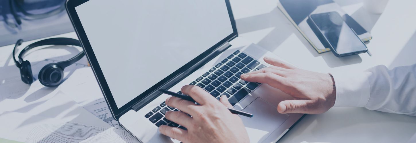A person typing on a laptop with a blank screen, holding a pen. Nearby are a smartphone, notebook, and headset on a bright, tidy desk. Professional setting.