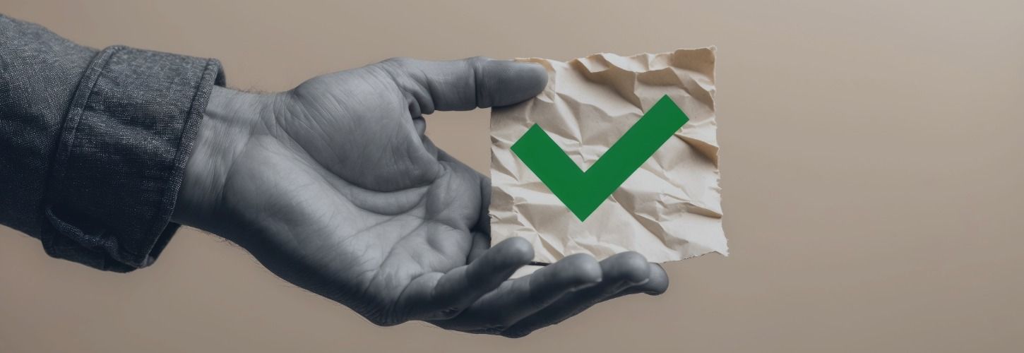 A grayscale hand holds a crumpled paper with a prominent green checkmark on it. The tone is positive, suggesting approval or success.