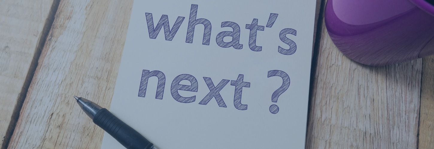 Notepad with "what’s next?" written on it, beside a pen and purple cup on a wooden table. Conveys curiosity and anticipation.