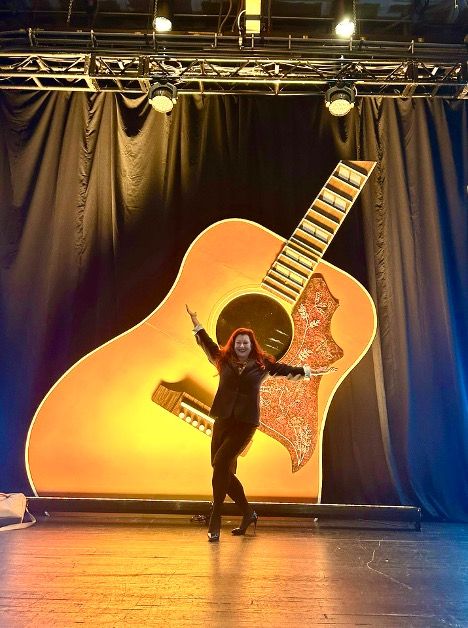 Kristy stands confidently on stage, arms spread wide, in front of a giant illuminated guitar backdrop. The setting is vibrant and celebratory.