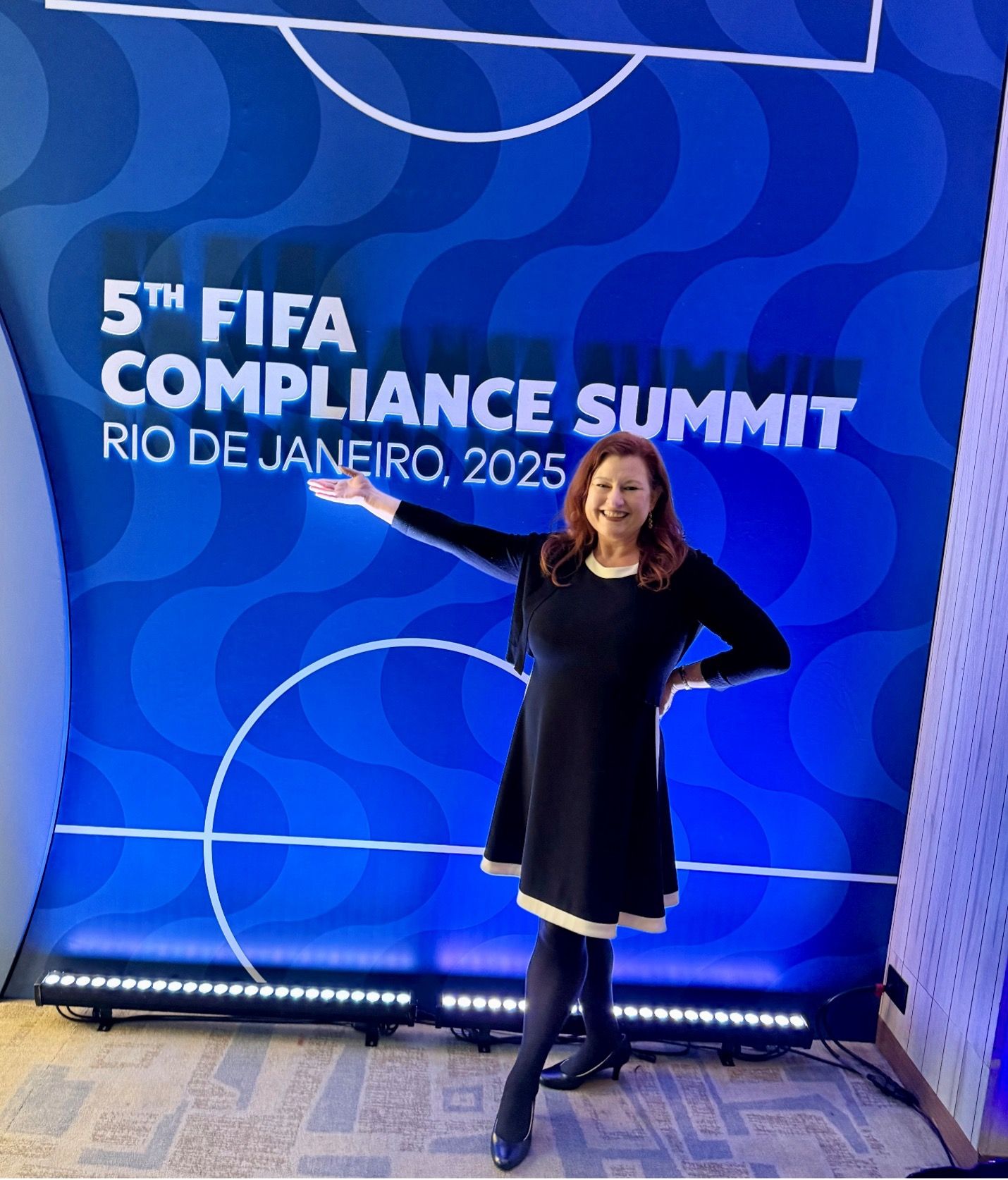 Kristy in a black dress with white trim stands smiling, gesturing towards a sign for the "5th FIFA Compliance Summit, Rio de Janeiro, 2025." The backdrop is vivid blue with white text.